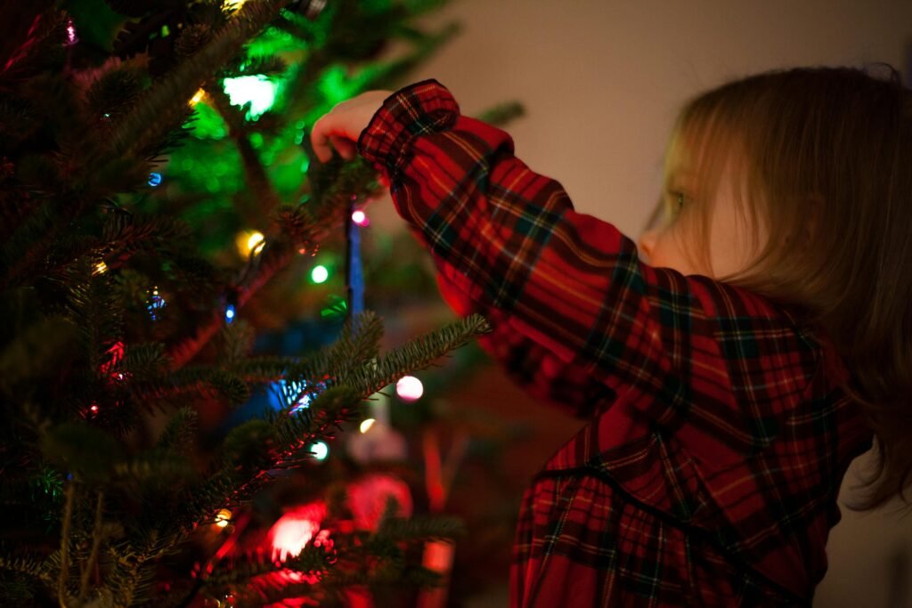 A young girl in plaid reaching to decorate a lit Christmas tree at home.